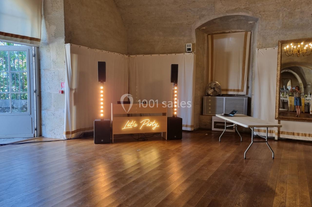 Salle avec parquet, table de mixage éclairée ’Let's Party’, enceintes et rideaux blancs, dans un cadre intérieur en pierre.