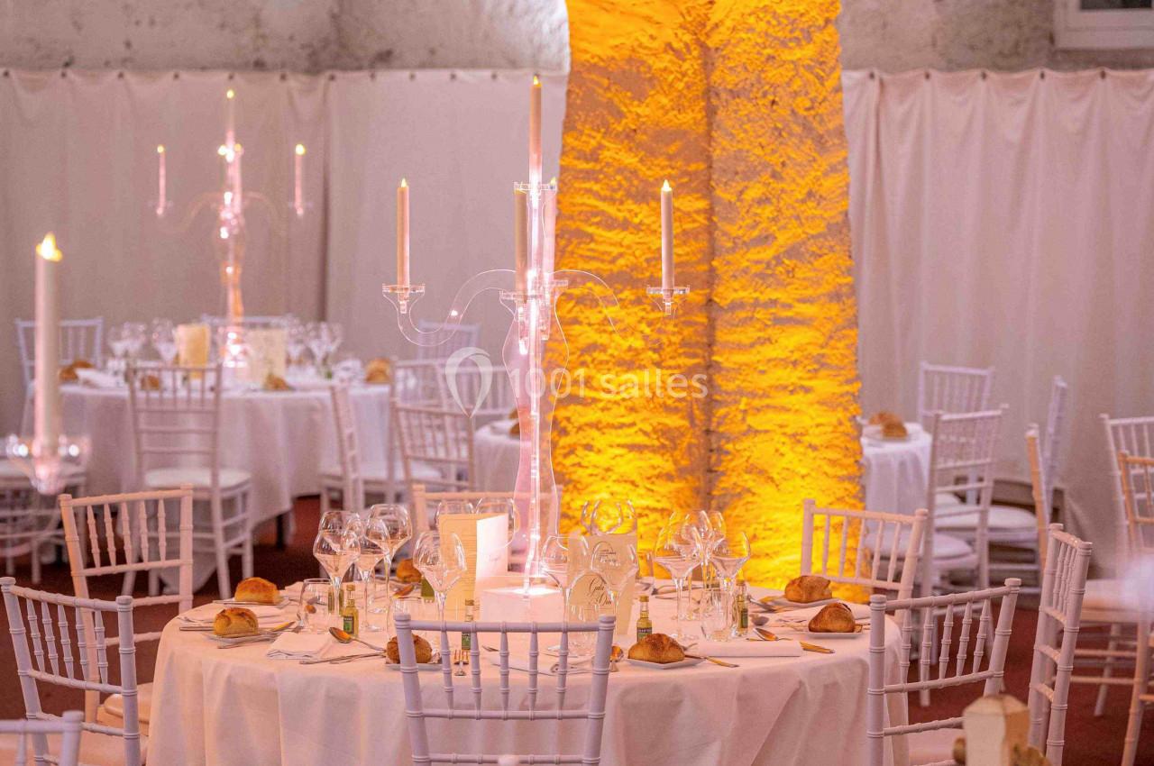 Salle voûtée décorée pour un événement, avec tables rondes dressées, chaises blanches et éclairage chaleureux.