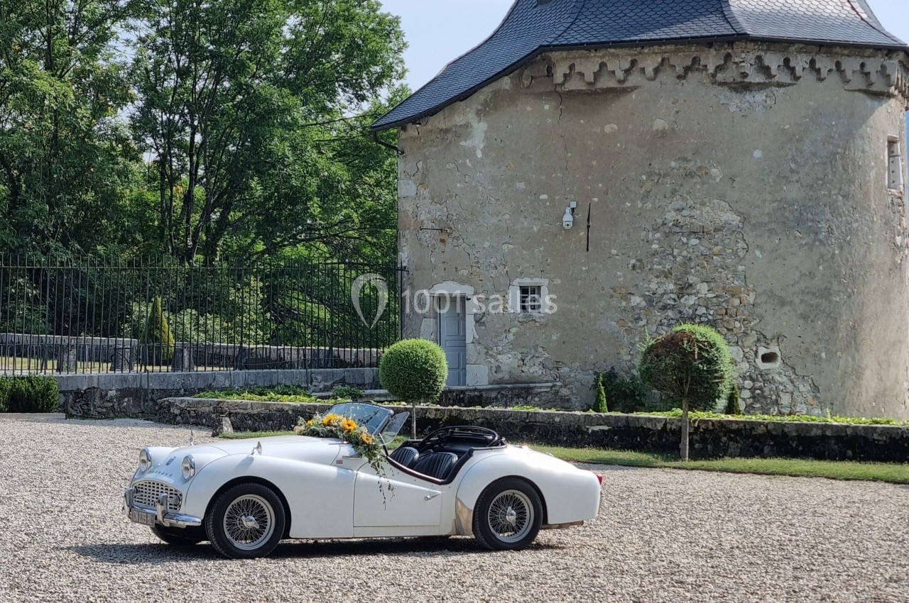 Voiture ancienne blanche décorée de fleurs, stationnée devant un bâtiment en pierre avec un toit en ardoise.
