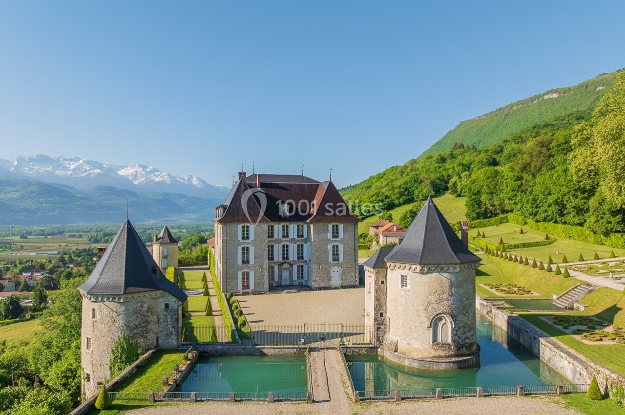Vue aérienne d'un château entouré de douves, avec des montagnes enneigées et une forêt en arrière-plan.
