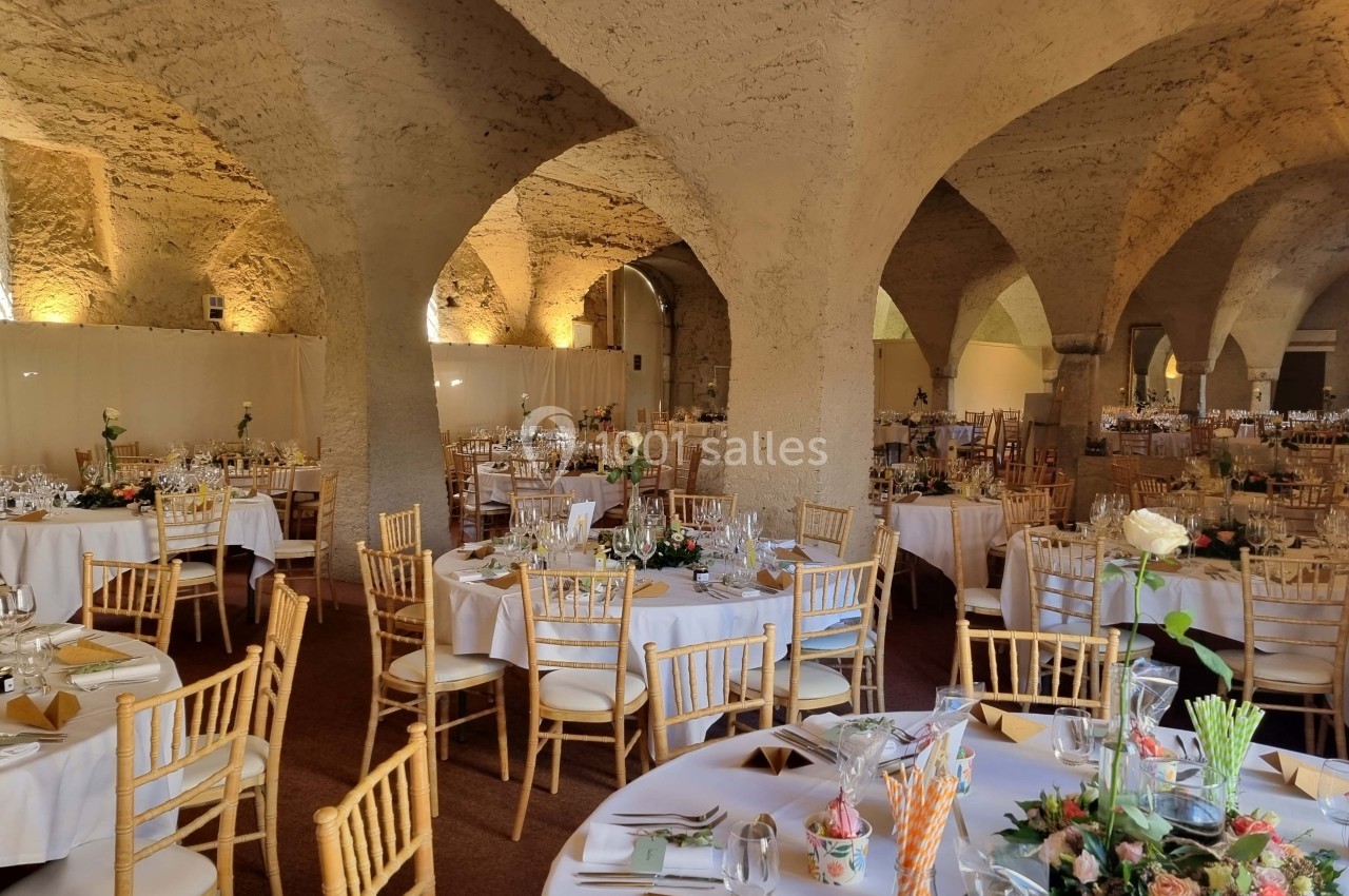 Salle voûtée décorée pour un événement, avec tables rondes dressées, chaises en bois clair et arrangements floraux.