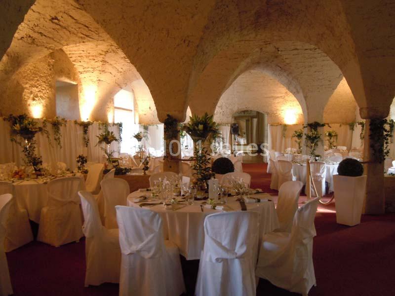 Salle voûtée décorée pour un événement, avec tables rondes dressées, nappes blanches et arrangements floraux.