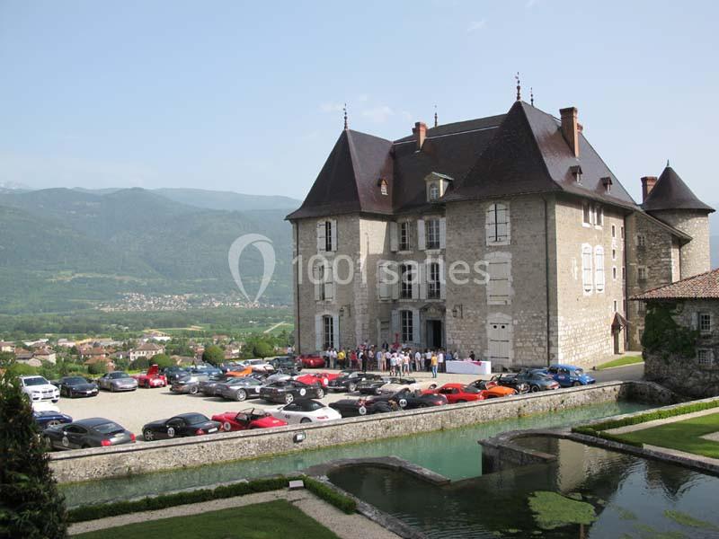 Château historique entouré de voitures de sport, avec un paysage de montagnes et de verdure en arrière-plan.