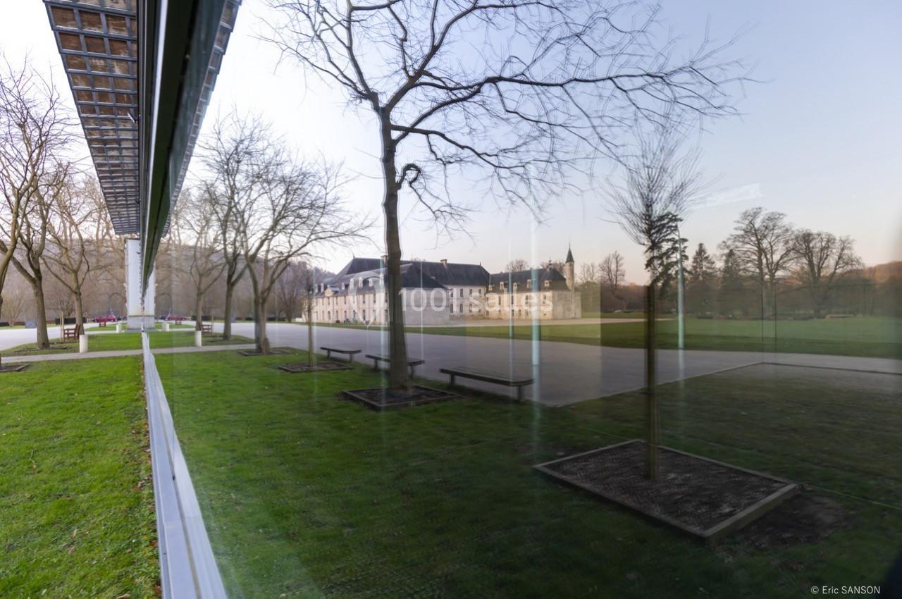 Reflet d'un bâtiment historique et d'arbres dans une large baie vitrée, avec pelouse et ciel dégagé.