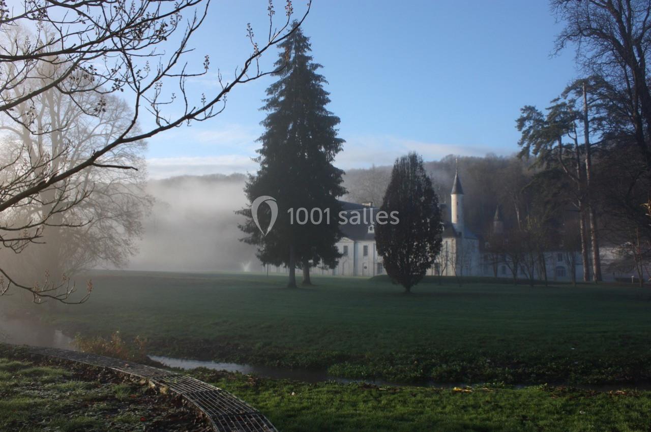 Un bâtiment ancien entouré d'arbres dans un parc verdoyant, avec de la brume matinale et un ruisseau au premier plan.