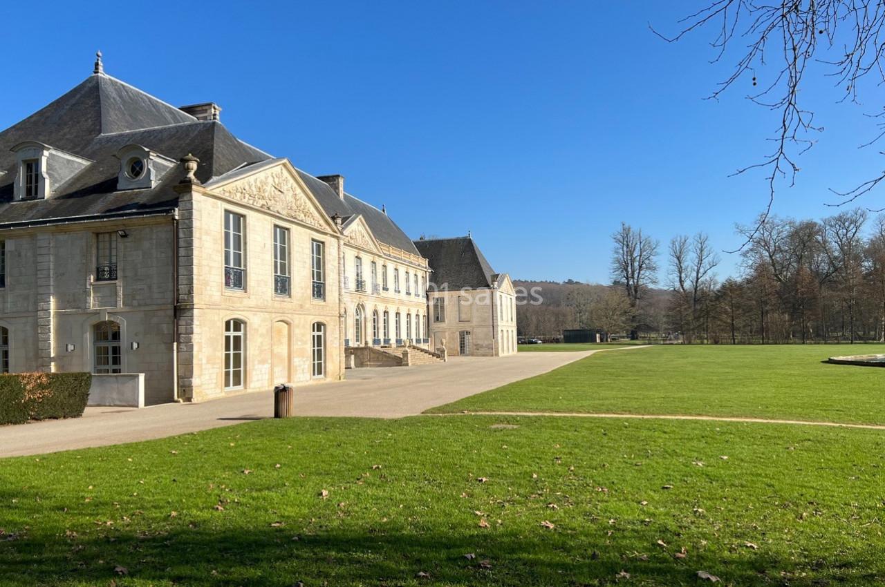 Bâtiment historique en pierre avec toit en ardoise, entouré d'une pelouse et d'arbres sous un ciel dégagé.