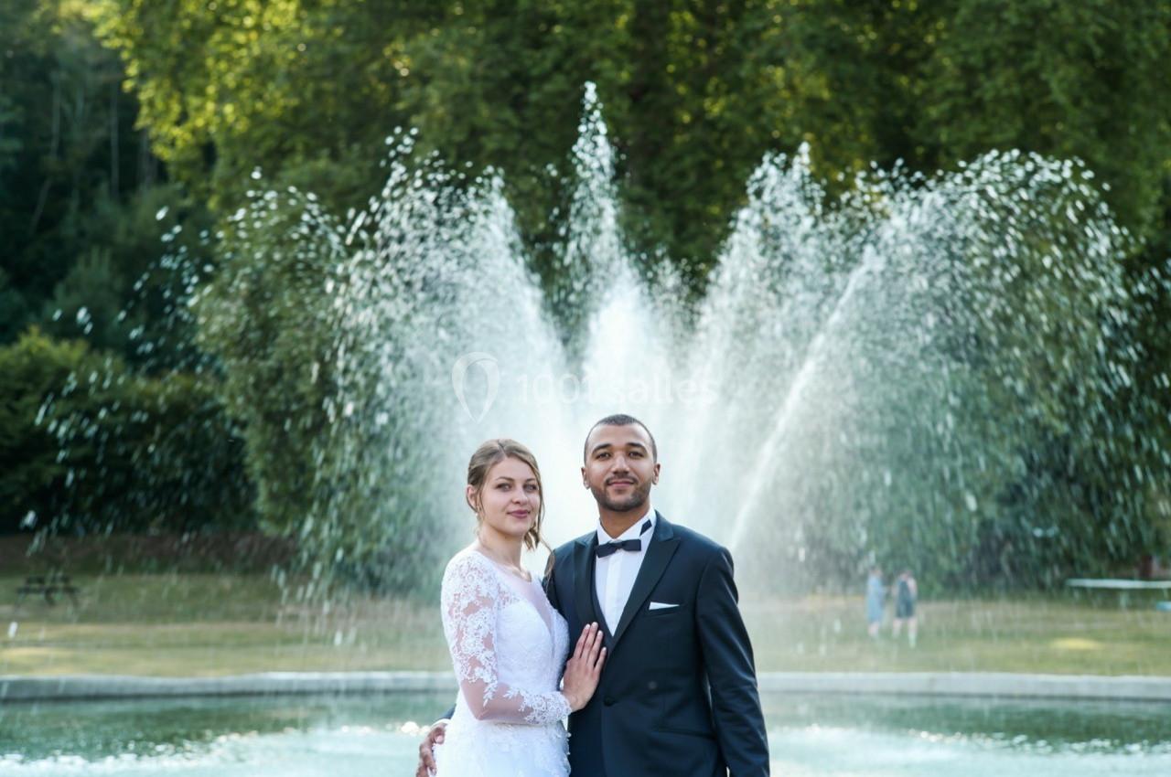 Un couple en tenue de mariage pose devant une fontaine jaillissante dans un parc verdoyant.