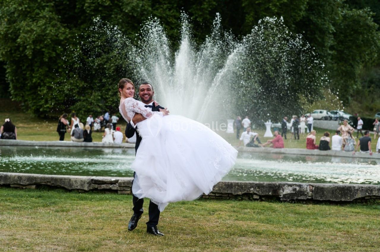 Un marié porte sa femme en robe blanche devant une fontaine, entourés d'invités dans un parc verdoyant.