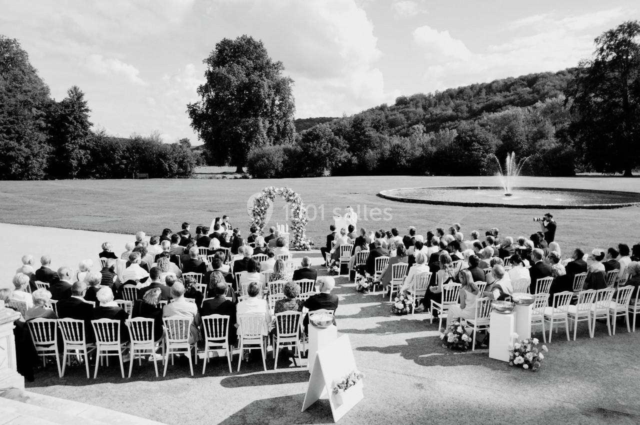 Cérémonie en plein air avec des invités assis devant une arche fleurie, près d'une fontaine et d'un parc verdoyant.