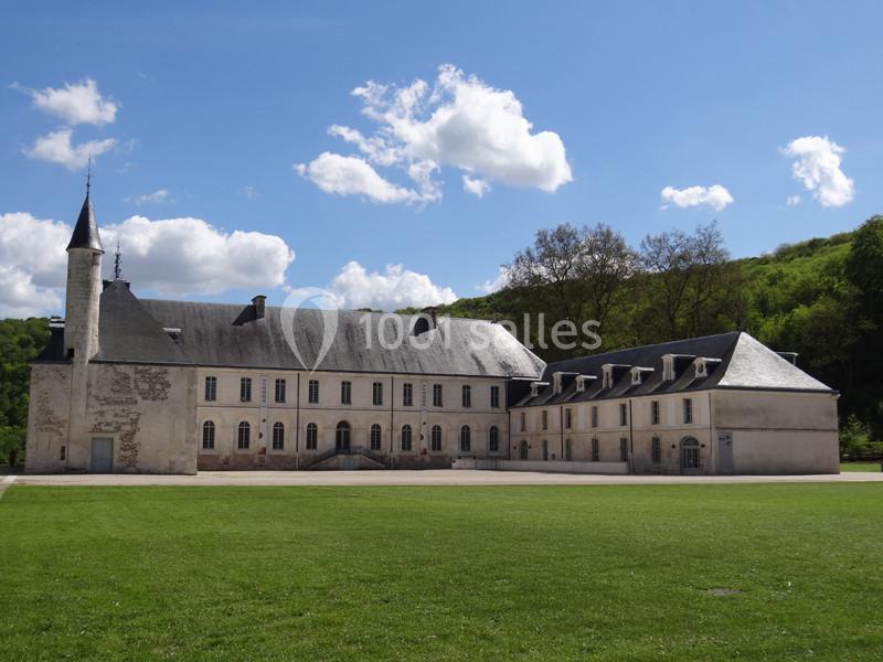 Bâtiment historique en pierre avec toit en ardoise, petite tour et grande pelouse au premier plan, sous un ciel bleu.