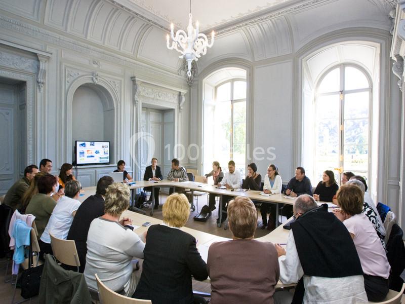 Réunion de personnes assises autour d'une grande table dans une salle lumineuse avec un écran affichant des présentations.