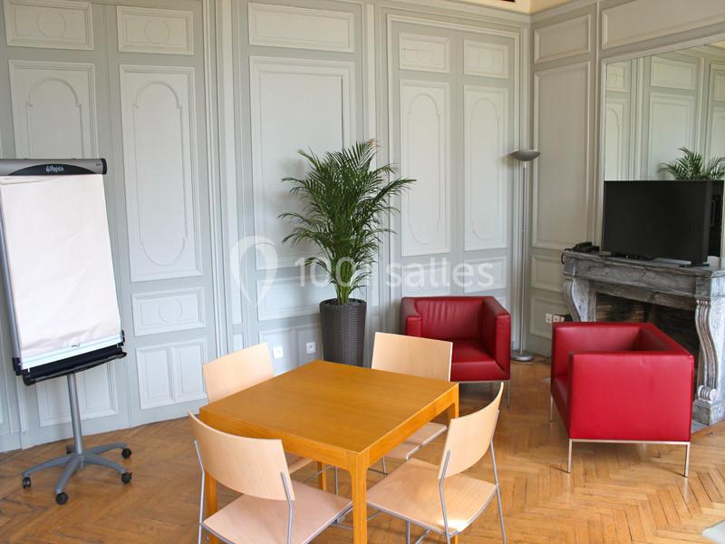 Salle lumineuse avec table en bois, chaises, fauteuils rouges, chevalet, plante verte et cheminée avec miroir.