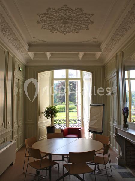 Salle lumineuse avec moulures au plafond, table ronde entourée de chaises, chevalet et vue sur un jardin par une grande…