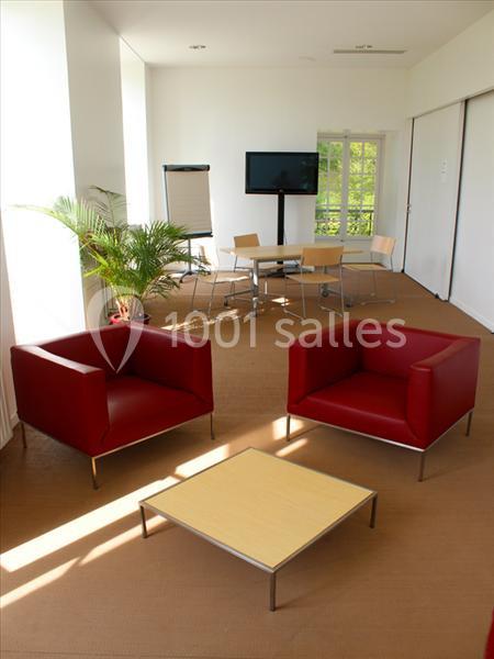 Salle lumineuse avec fauteuils rouges, table basse en bois clair, écran, chevalet et chaises près d'une fenêtre.