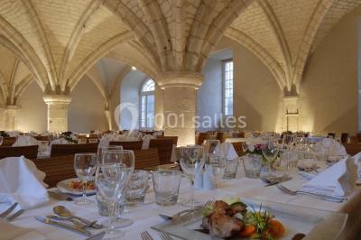 Salle voûtée élégamment dressée pour un repas, avec tables ornées de nappes blanches, verres et assiettes décorées.