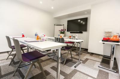 Salle de réunion aménagée en U avec tables, chaises colorées, écran, paperboard et bouteilles d'eau.