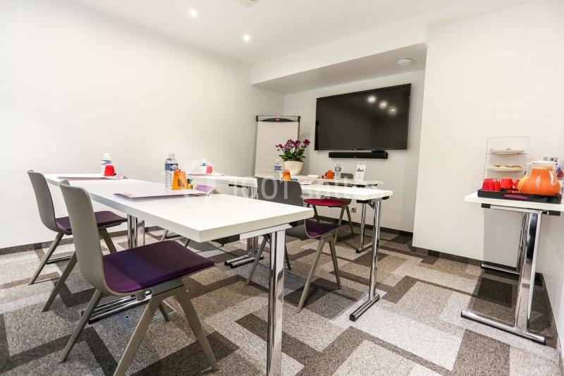 Salle de réunion lumineuse avec tables blanches, chaises violettes, écran mural, tableau blanc et boissons disposées.