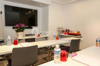 Salle de réunion aménagée en U avec tables, chaises colorées, écran, paperboard et bouteilles d'eau.