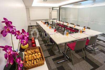 Salle de réunion aménagée en U avec tables, chaises colorées, écran, paperboard et bouteilles d'eau.