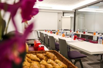 Salle de réunion aménagée en U avec tables, chaises colorées, écran, paperboard et bouteilles d'eau.