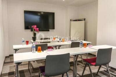 Salle de réunion aménagée en U avec tables, chaises colorées, écran, paperboard et bouteilles d'eau.