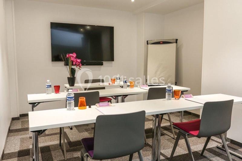 Salle de réunion équipée avec tables, chaises, écran, paperboard, bouteilles d'eau et décoration florale.