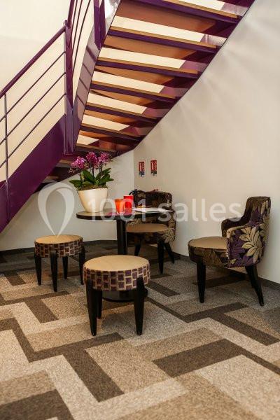 Espace avec table ronde, chaises et tabourets, sous un escalier violet, décoré d'une plante et de tasses rouges.