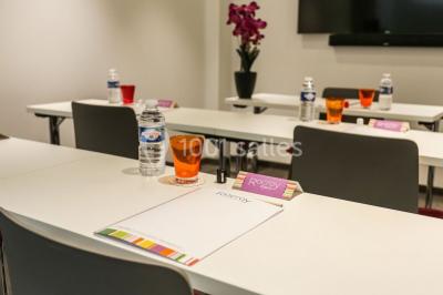 Salle de réunion aménagée en U avec tables, chaises colorées, écran, paperboard et bouteilles d'eau.