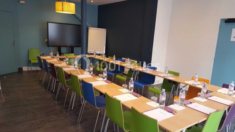 Salle de réunion équipée avec tables en U, chaises colorées, écran, paperboard, bouteilles d'eau et fournitures.