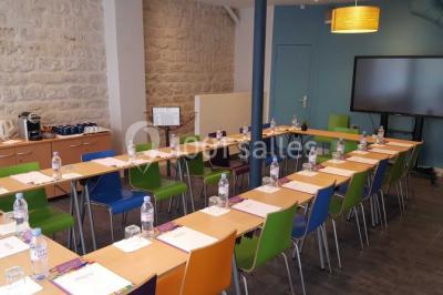 Salle de réunion aménagée en U avec tables, chaises colorées, écran, paperboard et bouteilles d'eau.