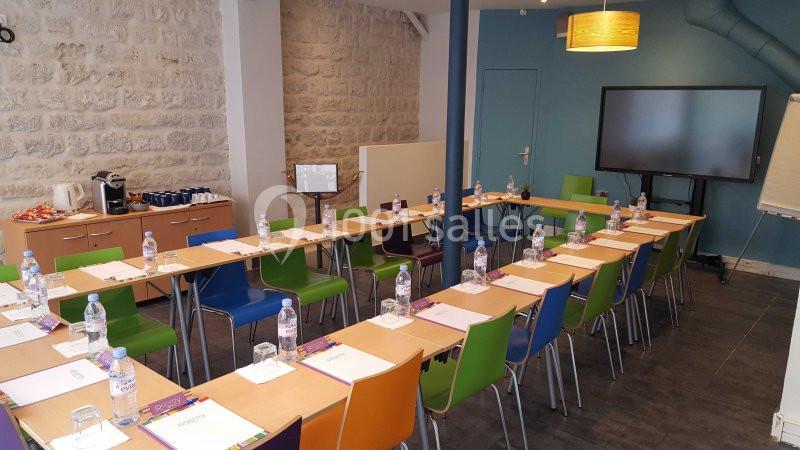 Salle de réunion aménagée en U avec tables, chaises colorées, écran, paperboard et bouteilles d'eau.