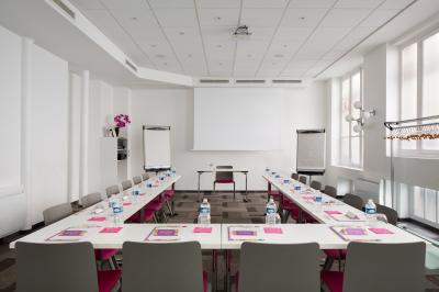Salle de réunion aménagée en U avec tables, chaises colorées, écran, paperboard et bouteilles d'eau.