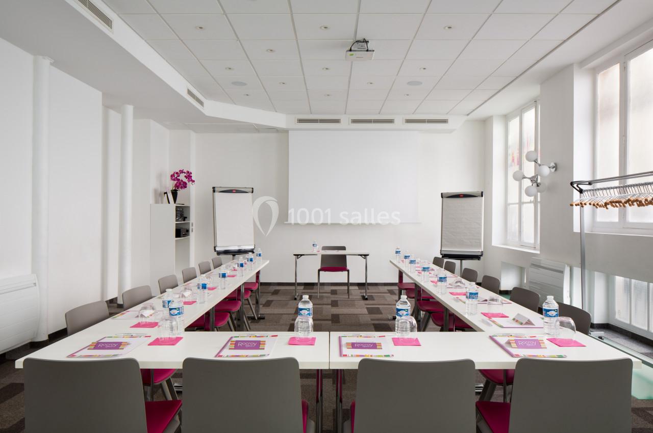 Salle de réunion lumineuse avec tables disposées en U, chaises, écran de projection et deux tableaux papier.
