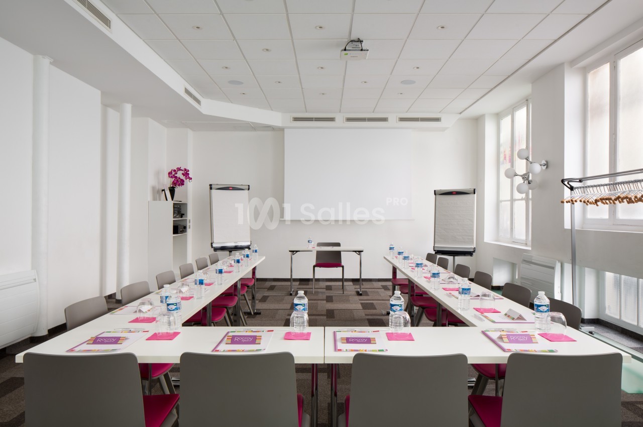 Salle de réunion lumineuse avec tables disposées en U, chaises, écran de projection et deux tableaux papier.