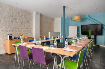 Salle de réunion aménagée en U avec tables, chaises colorées, écran, paperboard et bouteilles d'eau.