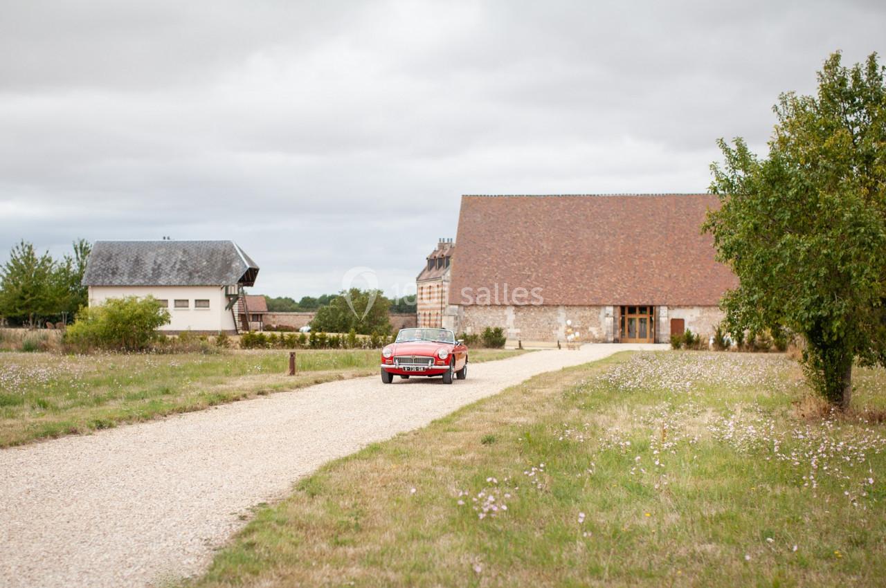 Une voiture rouge circule sur une allée gravillonnée devant des bâtiments ruraux et un arbre, sous un ciel nuageux.