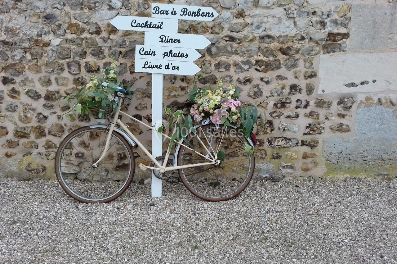 Un vélo décoré de fleurs est appuyé contre un panneau directionnel en bois devant un mur en pierre.