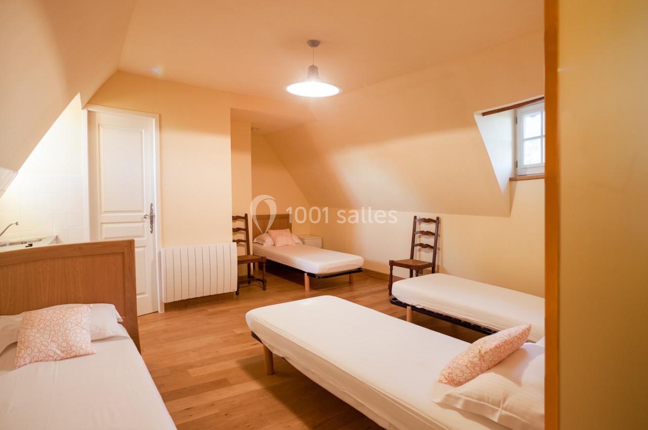 Chambre mansardée avec trois lits simples, parquet en bois, murs clairs et une petite fenêtre apportant de la lumière…