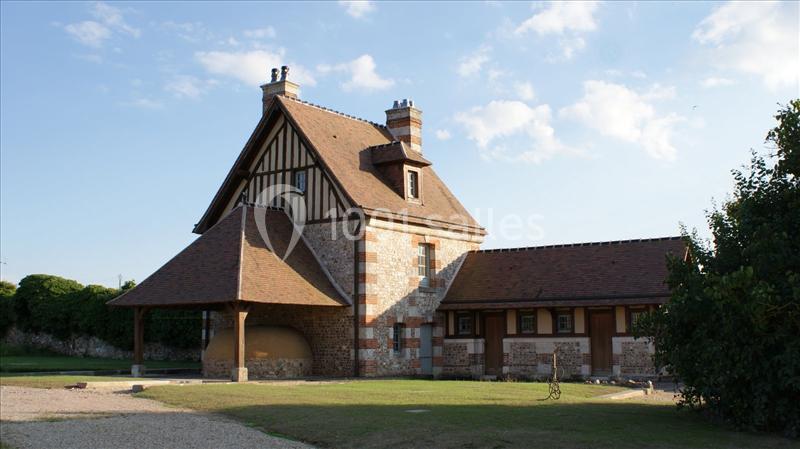 Maison à colombages avec toit en pente, entourée de pelouse et d'arbres, sous un ciel dégagé.