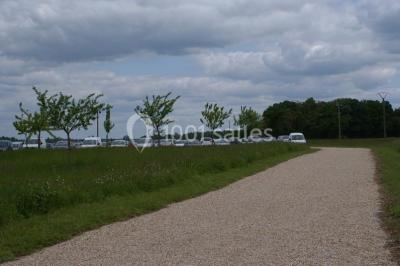 Chemin de gravier bordé d'herbe et d'arbres, avec des voitures garées en arrière-plan sous un ciel nuageux.