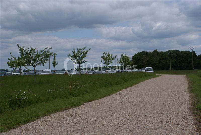 Chemin de gravier bordé d'herbe et d'arbres, avec des voitures garées en arrière-plan sous un ciel nuageux.