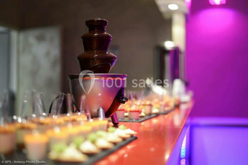 Fontaine à chocolat entourée de verrines colorées alignées sur une table dans un décor lumineux et moderne.