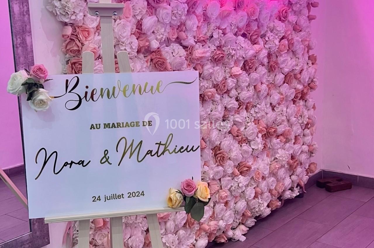 Mur de fleurs roses avec un panneau sur chevalet annonçant un mariage, éclairage néon en arrière-plan.