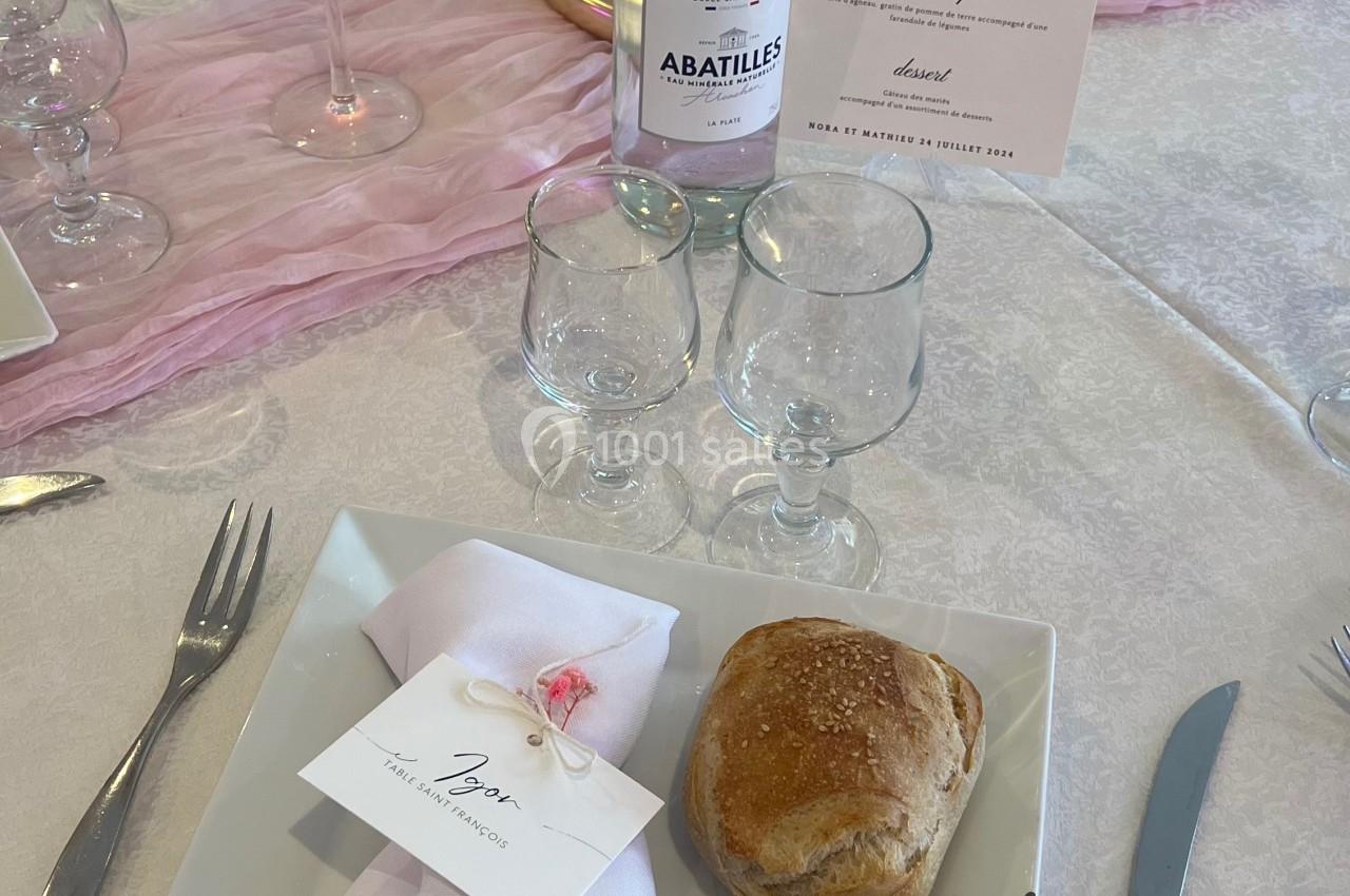 Table dressée avec une assiette carrée, serviette pliée, pain, menu, bougie, et bouteille d'eau minérale.