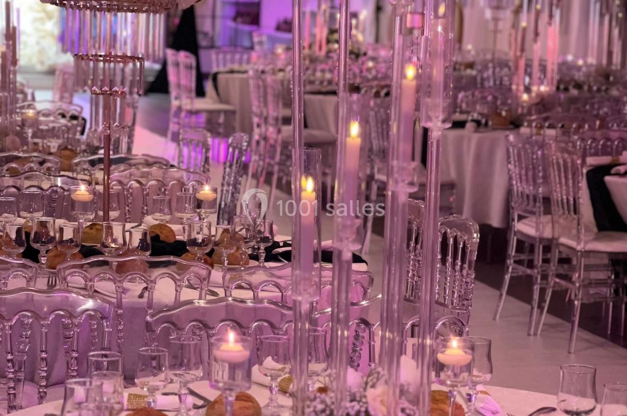 Salle de réception élégante avec tables dressées, chandeliers en verre, bougies allumées et décoration florale blanche.