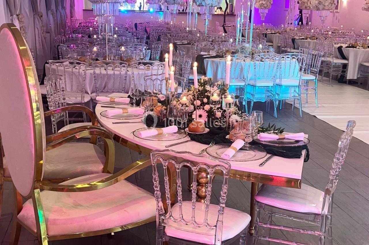 Salle de réception décorée avec des tables élégantes, des chandeliers et des chaises transparentes sous un éclairage coloré.