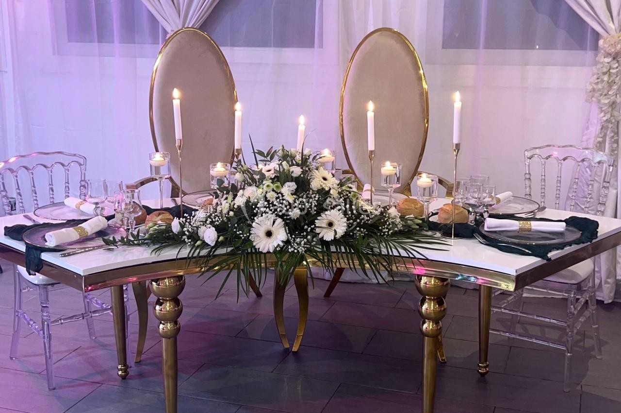 Table élégante décorée de fleurs blanches, bougies allumées et chaises transparentes, devant des rideaux clairs.