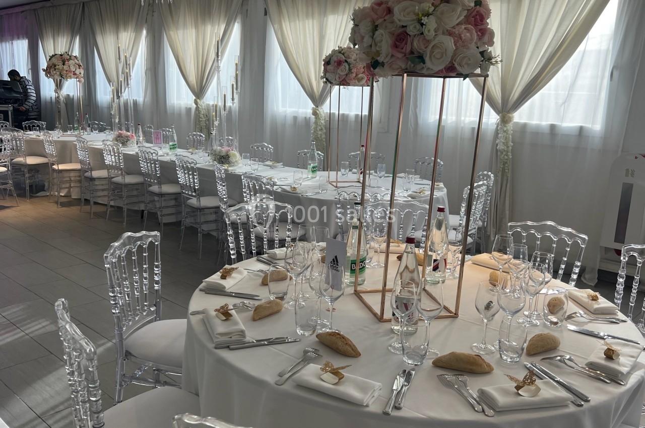 Salle de réception décorée avec des tables rondes dressées, chaises transparentes et centres de table fleuris.