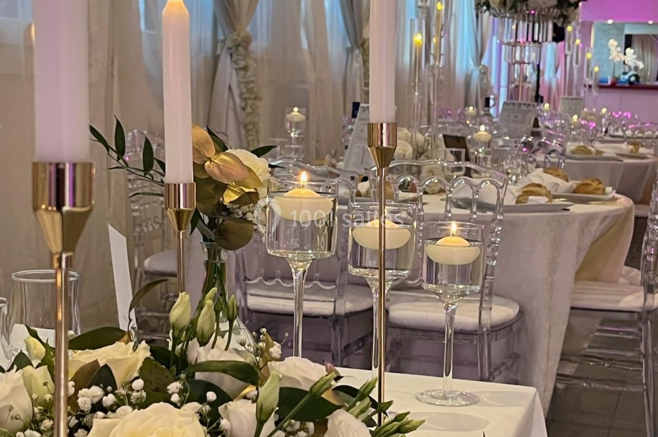 Salle de réception élégante avec tables décorées de fleurs blanches, bougies allumées et chaises transparentes.