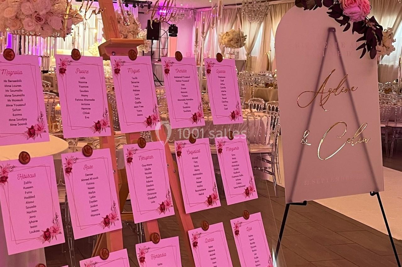 Plan de table de mariage avec des listes de noms sur un chevalet en bois et un panneau décoratif à côté, dans une salle…